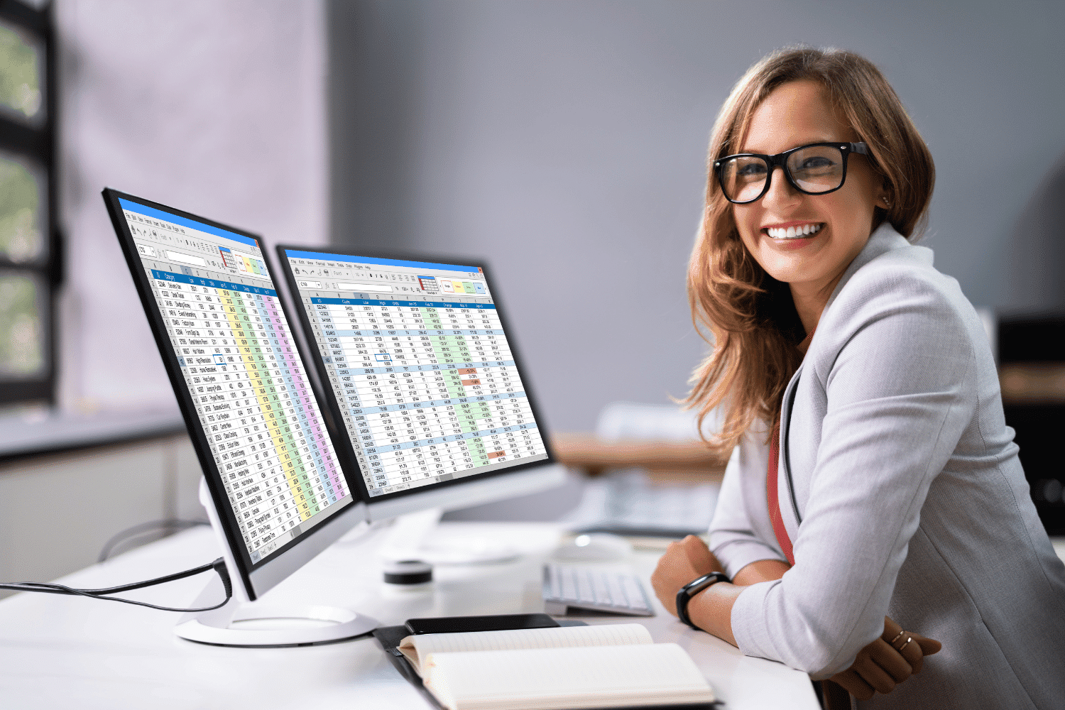 Young, smiling professional woman in front of dual-screen computer monitors with financial dashboard on screen