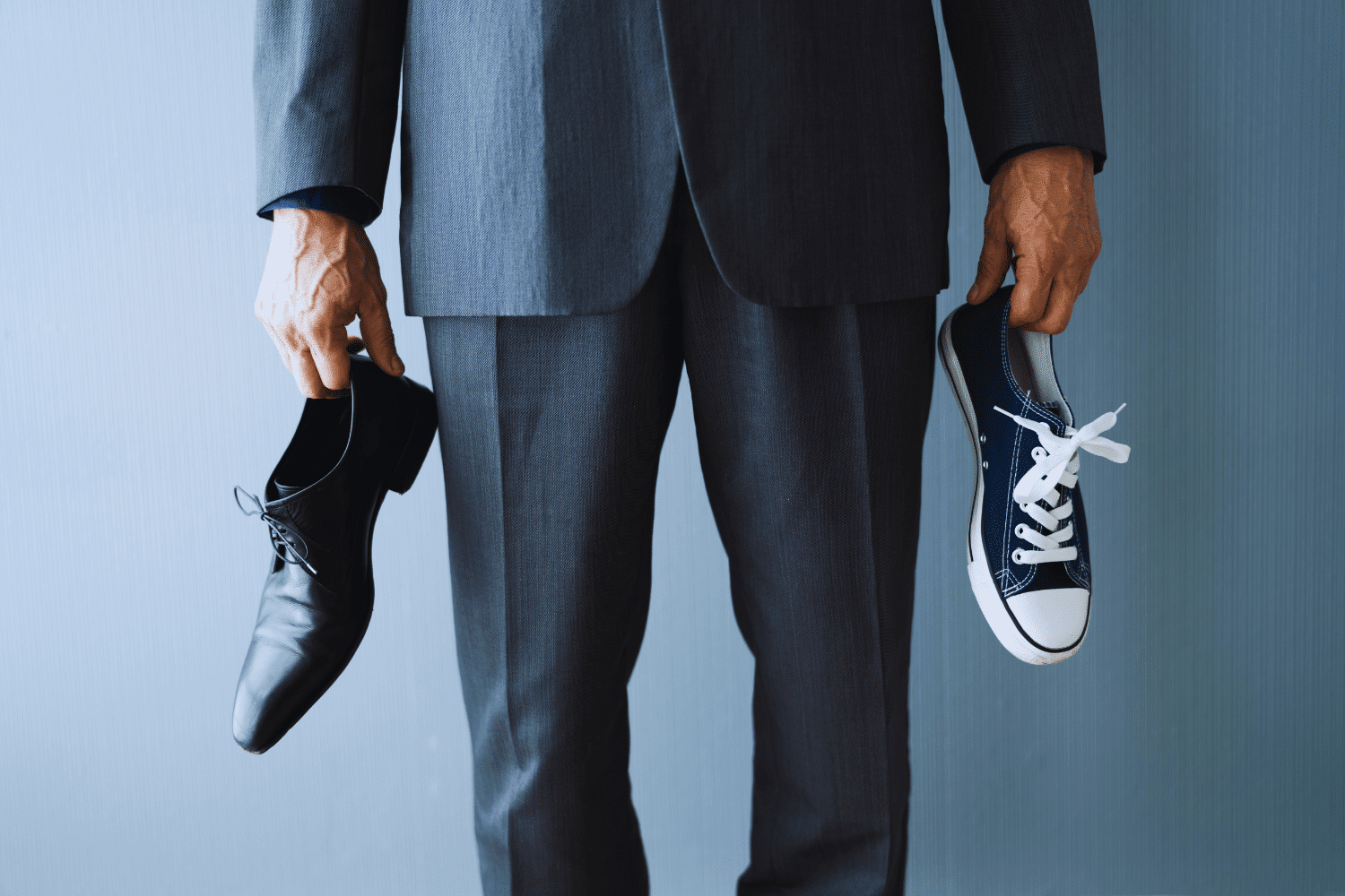 Man dressed in suit from waist down holding a dress shoe in one hand and a sneaker in the other