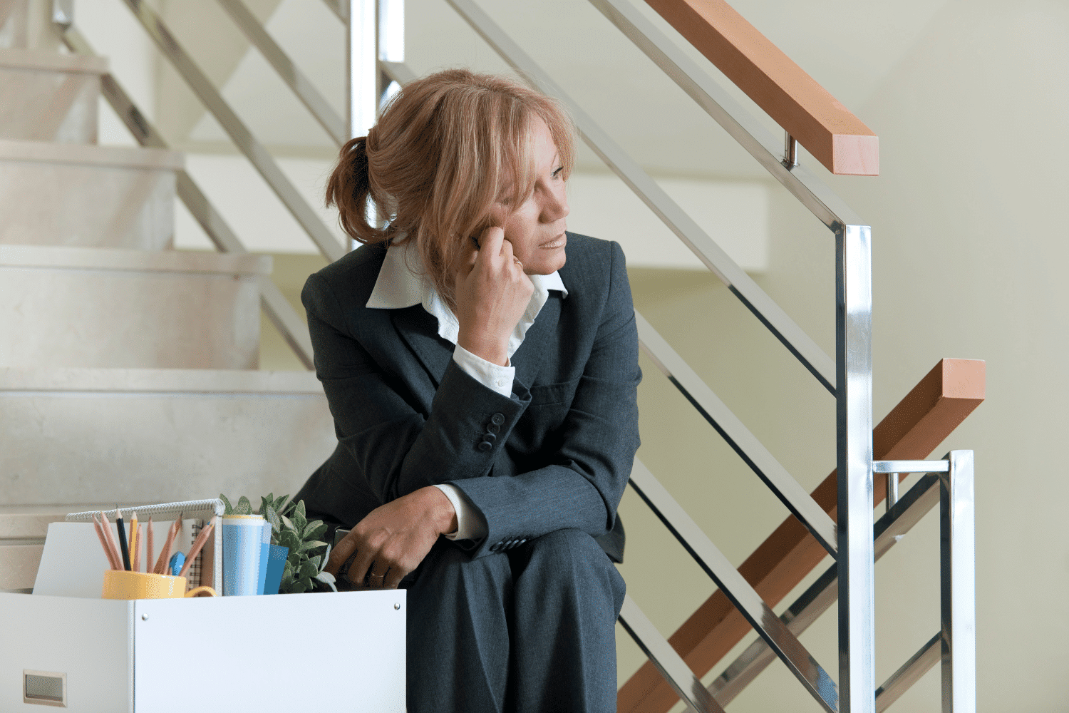 discouraged woman sitting on office stairs with box of belongings after being fired