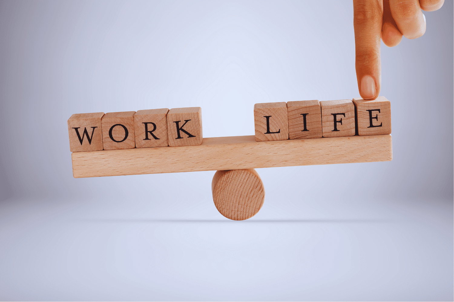 Scrabble tiles spelling WORK and LIFE balancing on a tray on a dowel with finger holding the LIFE side down