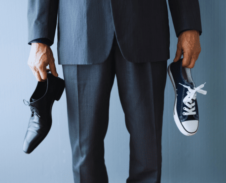 Man dressed in suit from waist down holding a dress shoe in one hand and a sneaker in the other