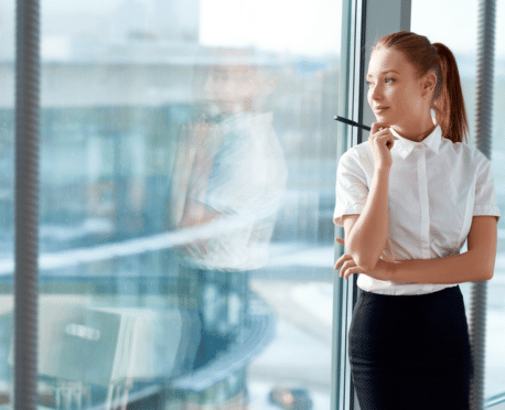 Young professional woman looking out office window, thinking about her dream job