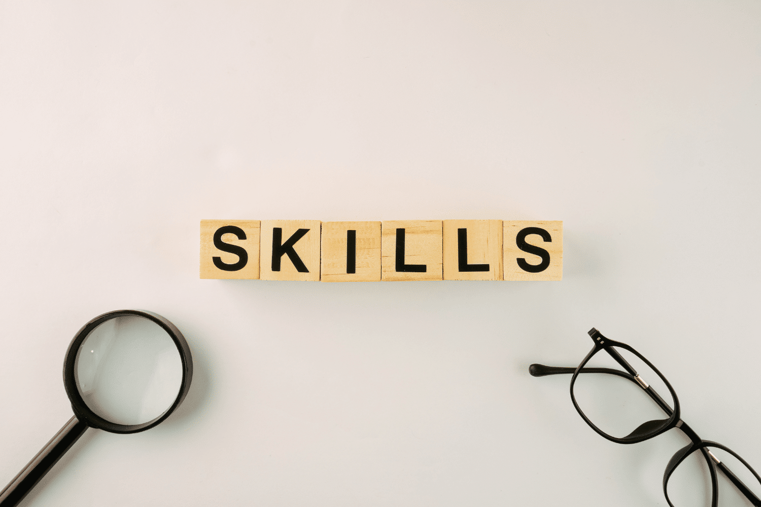 Overhead view of wooden letter blocks spelling SKILLS, flanked by a magnifying glass and glasses, symbolizing the detailed focus on candidate skills during executive search.