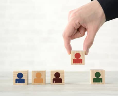 Selective executive hiring concept with hand choosing standout candidate block from diverse row of wooden people icons, symbolizing strategic leadership selection