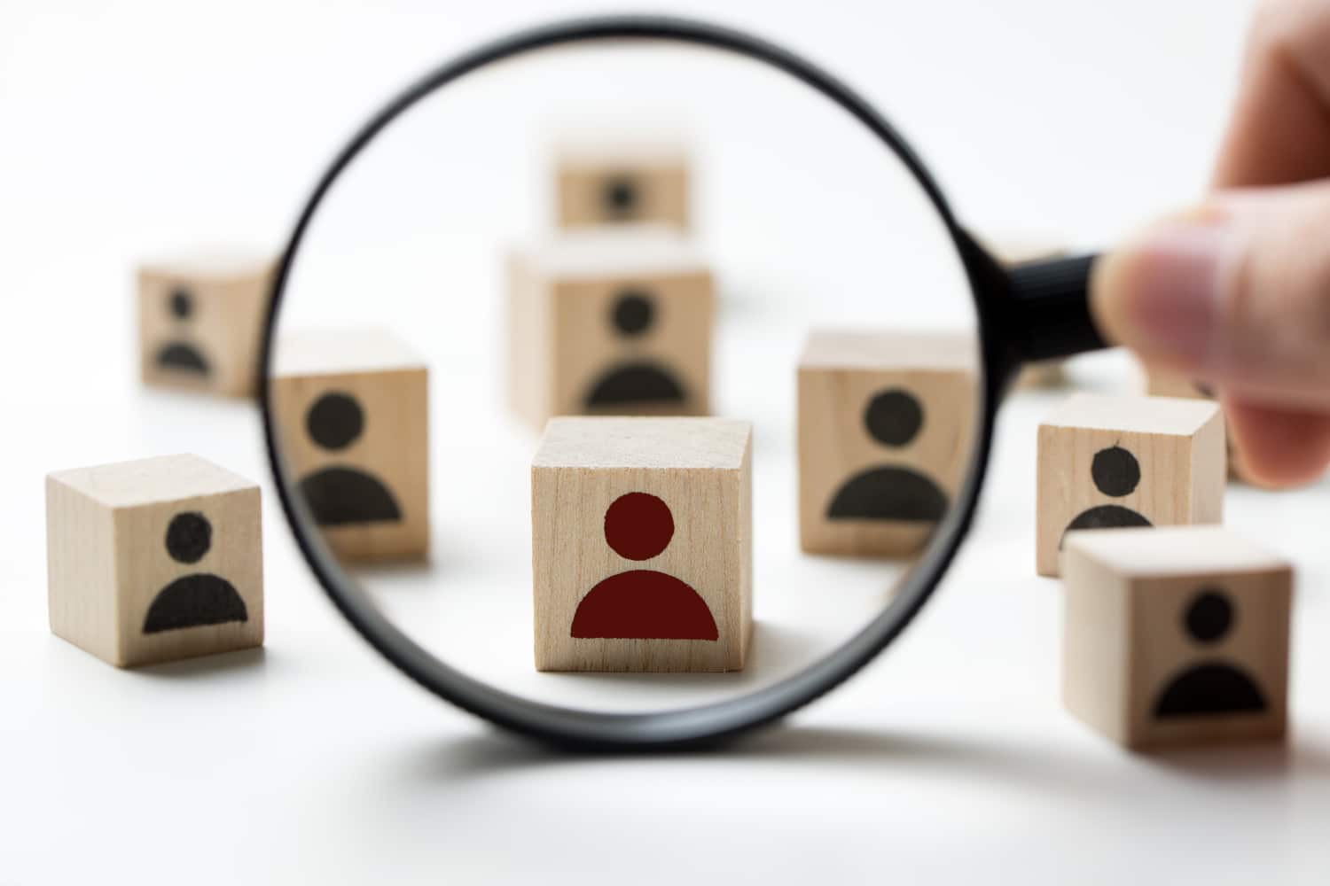 Hand holding a magnifying glass over wooden blocks with person icons, illustrating the research-heavy, data-driven process of identifying 