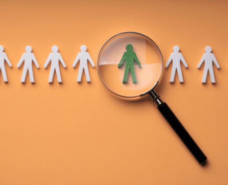 A magnifying glass identifies a distinct green candidate from a general row of white figures, symbolizing the challenges and solutions of CFO succession planning in 2026, and why specialized recruiters are key for Oggi Talent’s hiring strategy.