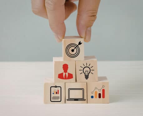 A conceptual visualization of building a succession-ready executive search capability. A hand strategically places a wooden block with a bullseye target atop a pyramid built from interlocking foundational elements: leadership capabilities (executive icon), innovation and strategy (lightbulb), and data-driven performance metrics (chart, laptop, growth graphs). This image illustrates the strategic, pro-active hiring reset necessary to create a competitive advantage.
